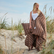 A woman standing on a sandy dune wrapped in brown jersey bedding with beige stripes, holding a matching pillow. Tall beach grass surrounds her under a blue sky.
